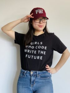 Vanessa is pictured with one hand on her head and she is wearing a maroon baseball hat that says MIT. She is also wearing a black t-shirt that says "Girls who code write the future" in capital, white letters, and jeans. Her other hand is on her hip.