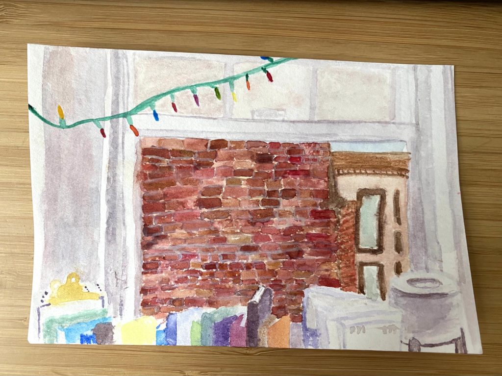 Watercolor painting of the view outside my bedroom window: a brick wall is outside, along with another brown building behind it. Inside, there is a line of books and a lamp, while Christmas lights dip in and out of the painting in the top left corner.